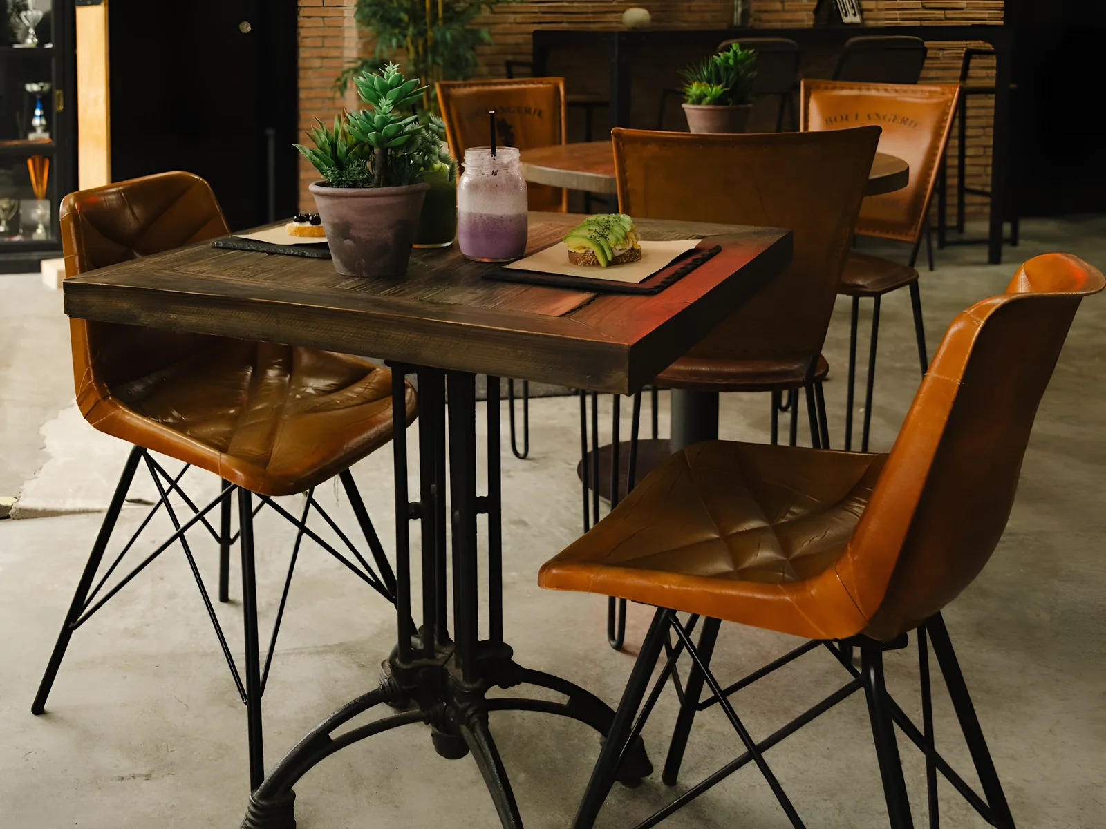 Mesa de madera con pie de hierro fundido y sillas de cuero coñac sobre el suelo de hormigón de La Calma, con un smoothie y una tosta